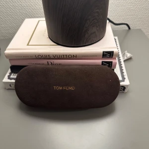 Brunt glasögonfodral från Tom Ford - Snyggt och stilrent glasögonfodral från Tom Ford i mörkbrunt mocka-material. Fodralet har en oval form och guldtext med loggan på ovansidan. Perfekt för att skydda dina glasögon med lite extra lyxig känsla.
