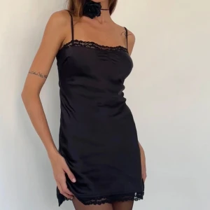 Svart kort klänning  - Supersnygg svart kort klänning från Motel i satin med tunna axelband och spetsdetaljer vid halsringning och nederkant. Klänningen har en slim passform och har justerbara axelband. Passar perfekt till fest och finare tillfällen. Oanvänd och med prislapp kvar.