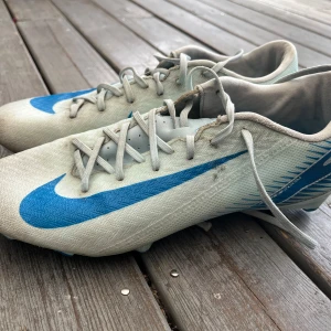 Nike fotbollsskor i vitt och blått - Säljer ett par Nike fotbollsskor i vitt med stora blå swoosh-loggor på sidorna. Skorna har snörning och är tillverkade i syntetmaterial med dobbar under sulan för bra grepp på planen. Perfekta för dig som vill sticka ut med stil på fotbollsplanen.