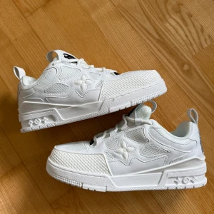 Louis Vuitton vita sneakers storlek-44 - Snygga vita Louis Vuitton sneakers med ikoniska LV-detaljer och blommönster på sidorna. Skorna har en chunky sula, snörning och coola logopatchar bak. Tillverkade i skinn och mesh för en lyxig och sportig vibe. Perfekta för dig som vill sticka ut med exklusiva sneakers.