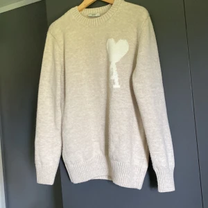Beige stickad tröja från Ami med hjärta - Säljer en beige stickad tröja från Ami med ett vitt hjärta och bokstaven A på bröstet. Tröjan har rund halsringning, ribbade muddar och lång ärm. Perfekt för dig som gillar stilrena och trendiga plagg med en twist.