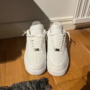 Nike Air Force 1 helvita sneakers - Klassiska Nike Air Force 1 sneakers i helvitt skinn med perforerad tå, rund tå och platt sula. Swoosh-logga på sidorna, broderad Nike Air på hälen och metallbricka med AF1 på snörningen. Perfekt clean look som alltid funkar.