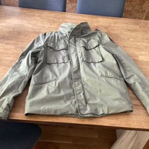 Grön vindjacka från Parajumpers - Snygg grön vindjacka från Parajumpers med klassisk patch på ärmen. Jackan har flera fickor framtill, hög krage och dragkedja. Tillverkad i polyester och perfekt för vår och höst. Stilsäker och funktionell jacka för dig som gillar streetwear.