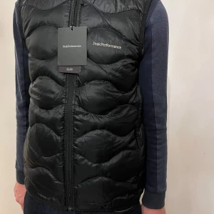 Svart dunväst från Peak Performance - Snygg svart dunväst från Peak Performance med vågformat quiltat mönster och dragkedja framtill. Västen har en stilren look med diskret logga på bröstet och är perfekt att slänga över en hoodie eller tröja för extra värme.