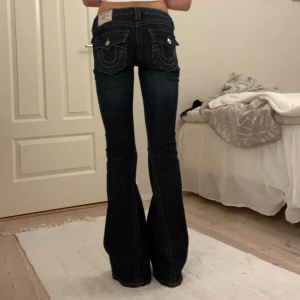 Mörkblå bootcut jeans från True Religion - Säljer ett par mörkblå bootcut jeans från True Religion med snygga kontrastsömmar och ikoniska fickdetaljer bak. Jeansen har låg midja, klassisk femficksdesign och är tillverkade i stretchig bomullsdenim. Perfekta för dig som gillar en retro vibe och markerad siluett. De är i fint skick men har satt ett snöre i midjan då de är lite för stora på mig och det är även därför jag säljer dem. Snöret syns inte inte direkt när man har på sig byxorna och går lätt att ta bort!💕 Kom gärna med bud🫶🏼