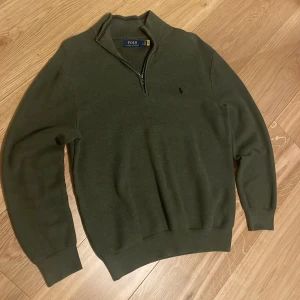 Olivgrön stickad tröja Polo Ralph Lauren - Snygg olivgrön stickad tröja från Polo Ralph Lauren med half zip och klassisk broderad logga på bröstet. Tröjan har lång ärm, ribbade muddar och en skön, normal passform. Perfekt för lager-på-lager och chill höst- och vinterstil.