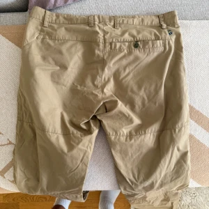 Beige shorts från Fjällräven G-1000 - Säljer ett par snygga beige shorts från Fjällräven i G-1000-material. De har praktiska fickor, bälteshällor och knappstängning. Perfekta för dig som gillar friluftsliv och vill ha slitstarka shorts med klassisk look och cool logga på benet.