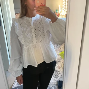 Vit blus med spets och volanger - Superfin vit blus med broderade spetsdetaljer och volanger på axlarna. Blusen har långärmade puffärmar och knappar framtill.