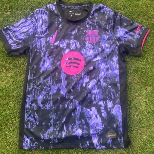 Barcelona x Nike lila/svart fotbollströja - Säljer en Barcelona fotbollströja från Nike i svart med lila och rosa detaljer. Tröjan har ett unikt abstrakt mönster, rosa Nike-logga och FCB-emblem samt Spotify-logga på bröstet. Rund hals och korta ärmar, tillverkad i lätt polyester som andas.