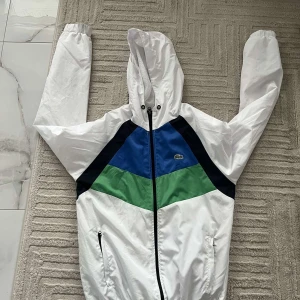 Lacoste vindjacka med färgblock S - Snygg vindjacka från Lacoste i vitt, blått, grönt och svart med klassisk krokodillogga på bröstet. Jackan har huva, dragkedja framtill och två fickor med dragkedja. Lätt meshfoder på insidan och sportig design med färgblock.