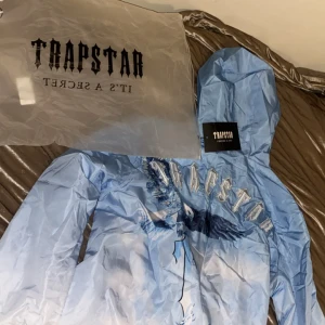 Blå vindjacka från Trapstar med tryck - Säljer en blå vindjacka från Trapstar i storlek M. Jackan har huva, dragkedja och ett stort tryck med fågelmotiv framtill. Lätt och smidig jacka med cool design och tydlig Trapstar-logga över bröstet. Perfekt för dig som vill sticka ut.