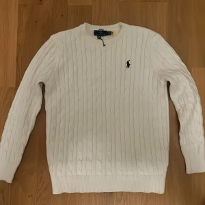 Vit kabelstickad tröja Polo Ralph Lauren - Vit kabelstickad tröja från Polo Ralph Lauren med klassisk rund halsringning och broderad marinblå logga på bröstet. Tröjan har lång ärm och ribbade muddar för en clean och tidlös look. Perfekt för lager-på-lager under kyliga dagar.