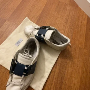 Valentino Open sneakers vit/blå - Snygga Valentino Open sneakers i vitt skinn med breda blåa band över vristen. Klassisk låg modell med rund tå och platt sula. Perfekta för dig som vill ha en stilren men ändå unik sneaker med exklusiv känsla.
