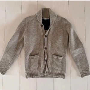 Beige stickad cardigan från J.Lindeberg - Snygg beige stickad kofta från J.Lindeberg med grovstickad struktur, sjalkrage och knappar framtill. Två fickor framtill och ribbade muddar ger en chill och klassisk vibe. Perfekt lager-på-lager-plagg för kyliga dagar. Storlek M passar även S