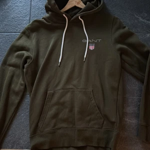 Olivgrön hoodie från GANT med logga - Snygg olivgrön hoodie från GANT med vit broderad logga och emblem på bröstet. Tröjan har huva med vita snören, stor magficka och ribbade muddar. Perfekt för chill dagar och enkel att matcha med jeans eller joggers.