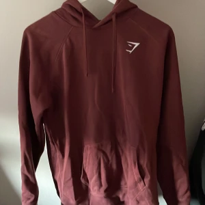 Vinröd hoodie från Gymshark - Säljer en vinröd hoodie från Gymshark i storlek M. Tröjan har huva med snörning, en stor magficka och långärmade ärmar. Perfekt för chill eller träning. Diskret Gymshark-logga på bröstet. Materialet är mjukt och känns skönt mot huden.