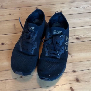 Svarta EA7 Emporio Armani sneakers - Svarta sneakers från EA7 Emporio Armani med guldiga logodetaljer på sidan. Skorna har stickad ovandel i syntetmaterial, snörning och platt sula. Perfekta för dig som gillar stilrena och sportiga skor med exklusiv känsla.