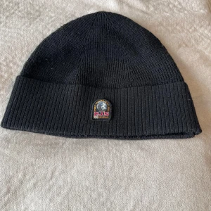 Svart ribbad mössa Parajumpers - Svart ribbstickad mössa från Parajumpers med uppvikt kant och snyggt broderat märke framtill. Klassisk och enkel design som passar till allt, tillverkad i mjuk ull som håller dig varm under kalla dagar. Storlek S, M