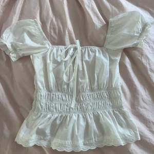 Vit blus från PrettyLittleThing - Supersöt vit blus med puffärmar och smockad midja från PrettyLittleThing. Toppen har  volangkant nertill och spetsdetaljer längs ärmslut och nederkant. 