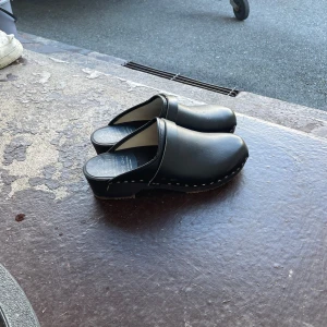 Svarta trätofflor i skinn - Säljer ett par klassiska svarta trätofflor från Troentorp med ovandel i skinn och sula i trä. Modellen har rund tå och en diskret klack, med vita sömmar längs kanten för en snygg kontrast. Perfekt för dig som gillar enkel och tidlös stil.