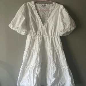 Vit broderad sommarklänning S - Superfin vit klänning med broderade blommor och v-ringning. Puffiga korta ärmar och volangiga lager ger en romantisk vibe. Perfekt för varma dagar och har en luftig passform. Tillverkad i bomull för en skön känsla.
