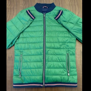 Grön pufferjacka med blå detaljer - Stilren grön pufferjacka med marinblå muddar och dragkedja framtill. Jackan har nästan aldrig kommit till användning. Den är tunnare och perfekt inför sen sommar och höst! Är ni frågor så tveka inte att hojta till! Pris går alltid att diskutera.