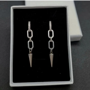 Set med silverfärgade örhängen och halsband - Säljer ett set med coola silverfärgade örhängen med kedjelänkar och spetsiga hängen samt ett matchande halsband med en kedja och en hängande vit pärldroppe formad som en ängel. Perfekt för dig som gillar stilrena och trendiga accessoarer.