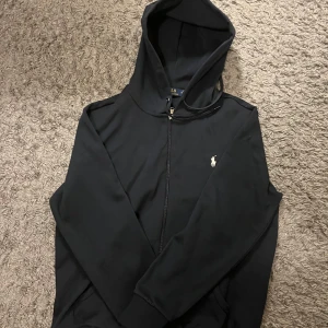 Svart hoodie från Ralph Lauren  - Svart hoodie från Ralph Lauren med dragkedja och klassisk broderad logga på bröstet. Tröjan har huva, långa ärmar och känguruficka framtill. Tillverkad i mjuk bomullsmix som är skön att bära. Perfekt för en chill och stilren look.