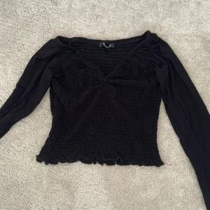 Svart  topp från Gina Tricot - Supersnygg svart långärmad topp från Gina Tricot med smockad struktur och v-ringning. Toppen har en figurnära passform och lätt volangkant nertill. Perfekt att styla med jeans eller kjol för en trendig look.