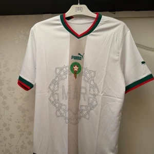 Marocko landslagströja Puma vit - Säljer en officiell Marocko landslagströja från Puma i vitt med gröna och röda detaljer på krage och ärmslut. Tröjan har korta ärmar, broderat landslagsemblem och subtilt mönster framtill. Perfekt för dig som vill stötta Marockos fotbollslandslag.