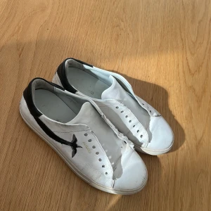Vit-svarta sneakers från Axel Arigato - Snygga vita sneakers från Axel Arigato med svart fågelmotiv på sidan och svart häl. Skorna har en klassisk låg profil med rund tå och platt sula. Guldtext med logga på sidan ger en clean och stilren look. Har nya snören men dom är inte på än