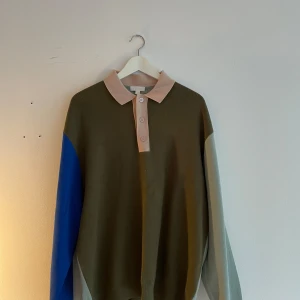 Färgstark pikétröja från COS i L - Säljer en unik pikétröja från COS i storlek L. Tröjan har en cool färgblockad design med olivgrön kropp, ljusblå och blå ärmar samt beige krage och knappslå. Långärmad och tillverkad i mjuk bomull, perfekt för dig som gillar stilrena men ändå lekfulla plagg.
