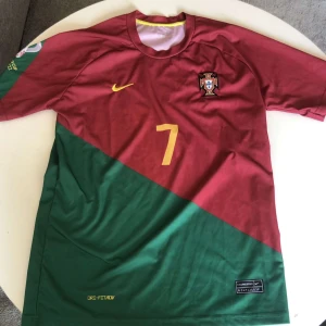 Portugal Ronaldo 7 fotbollströja Nike - Snygg Portugal fotbollströja, Nike med Ronaldo och nummer 7 i gult på ryggen och bröstet. Tröjan har en diagonal design i vinrött och grönt, med korta ärmar och rund hals. Materialet är lätt och andas, perfekt för match eller träning. Ikoniskt landslagsmärke på bröstet.