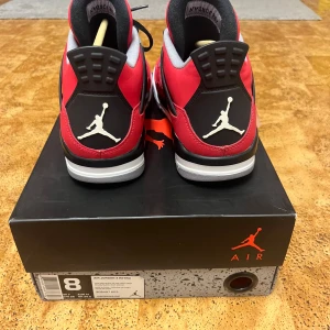 Nike Air Jordan 4 Retro toro bravo - Säljer ett par Nike Air Jordan 4 Retro toro bravo med ikonisk Jumpman-logga på hälen. Skorna har röd ovandel i mocka, svarta och grå detaljer, svart snörning och vit sula. Grå meshpaneler och klassisk siluett gör dem till ett måste för sneakerheads.