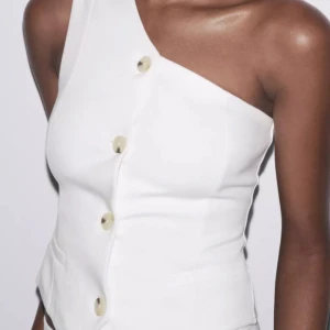 Vit asymmetrisk väst med knappar - Snygg vit väst med en axel och stora dekorativa knappar framtill. Västen har en stilren, figurnära passform och asymmetrisk skärning som ger en modern vibe. Perfekt att styla med både jeans och kostymbyxor för en trendig look.