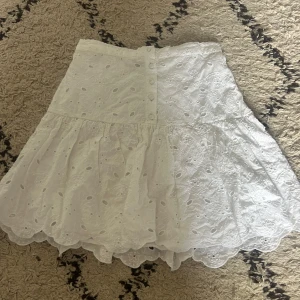 Vit broderad kortkjol med knappar - Superfin vit kortkjol med broderade blommor och snygga knappar framtill. Kjolen har volangkant och ett hålmönster som ger en somrig och fräsch vibe. Säljer pågrund av att den inte kommer till användning 