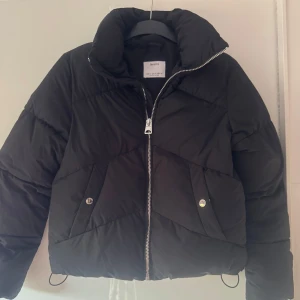 Ny. Svart pufferjacka från Bershka XS - Svart pufferjacka från Bershka i storlek XS. Jackan har hög krage, dragkedja framtill och två fickor med tryckknappar. Snygg quiltad design och en lite boxig passform som ger en cool vibe. Perfekt för kalla dagar när du vill vara både varm och stilren.