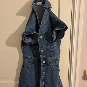 Kort jeansklänning med knappar - Snygg kort jeansklänning i blå denim med knappar framtill och korta ärmar. Klänningen har krage, bröstfickor och markerad midja. Perfekt för en cool och avslappnad stil.