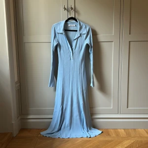 Ljusblå långklänning från Adoore - Supersnygg ljusblå långklänning från Adoore med ribbad struktur och klassisk krage. Klänningen har lång ärm, v-ringning med knappar framtill och är figurnära i passformen. 