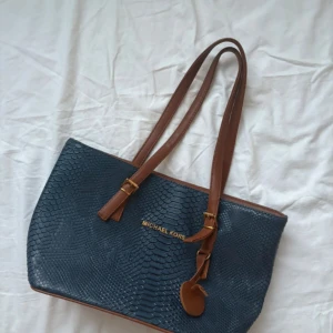 Blå  axelväska med ormmönster - Snygg axelväska  i mörkblått med ormmönstrad struktur och bruna handtag. Väskan har guldfärgade detaljer och flera fack inuti med dragkedja. Perfekt storlek för allt du behöver ha med dig och riktigt trendig look.