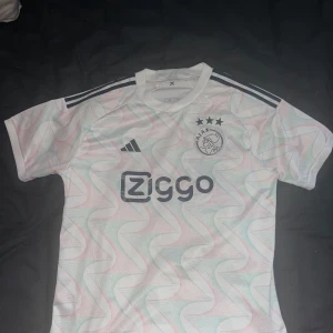 Ajax vit kortärmad fotbollströja Adidas - Ajax Amsterdam fotbollströja från 2023-2024 säsongen. Adidas i vit med pastellfärgade vågmönster i rosa och grönt. Kortärmad modell med rund hals, svarta Adidas-ränder på axlarna och Ziggo-logga på bröstet. Materialet är lätt och andas, perfekt för match eller träning.