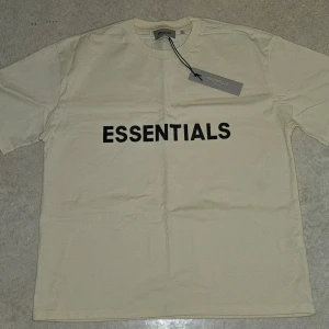 Essentials beige t-shirt i bomull - Beige t-shirt från Essentials med svart tryck över bröstet. Klassisk rund halsringning och korta ärmar. Tillverkad i mjuk bomull för en skön känsla. Perfekt för en clean och stilren look. Storleken är M men passformen är L (De är lite baggy) Helt oanvänd och taggen sitter kvar