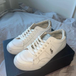 Vita sneakers från Polo Ralph Lauren - Snygga vita sneakers från Polo Ralph Lauren med silverglittriga detaljer på plösen och hälen. Klassisk låg modell med snörning och rund tå. Skorna har Polo-logga på sidan och med platt sula. Perfekta för en clean och fräsch look. Skriv hjärna prisförslag!