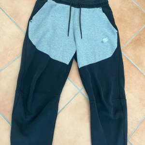 Nike tech byxor - Snygga mjukisbyxor från Nike i svart och ljusgrått med dragsko i midjan och stora fickor framtill. Byxorna har en sportig look och är perfekta för chill eller träning. Nike-logga på vänster sida. Storlek S