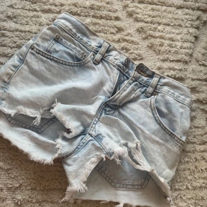 Ljusblå jeansshorts med fransig kant - Säljer ett par ljusblå jeansshorts med låg midja och fransiga, slitna kanter. Klassisk femficksmodell med dragkedja och knapp framtill. Perfekt för dig som gillar en chill och avslappnad stil på sommaren.