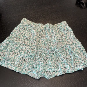 Blommig kortkjol med snörning - Supersöt kortkjol med blommigt mönster i vitt, svart och mintgrönt. Kjolen har volangig känsla och smal snörning i midjan för extra detalj. Perfekt för dig som gillar en luftig och lätt stil under varmare dagar. Har även under shorts.