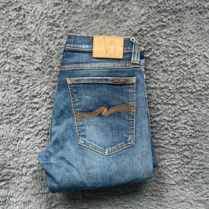 Blå jeans från Nudie Jeans - Säljer ett par klassiska blå jeans från Nudie Jeans med snygga kontrastsömmar och ikonisk broderad bakficka. Modellen har raka ben och normal passform, perfekt för dig som gillar stilrena jeans med en modern touch. Skicket är 9/10 utan defekter