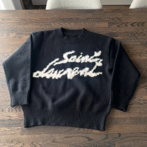 Svart stickad Saint Laurent tröja - Svart stickad tröja från Saint Laurent med vit fuzzy logga på bröstet. Tröjan har rund hals, ribbade muddar och är långärmad. Perfekt för dig som vill ha en statement piece med lyxig vibe och skön passform.