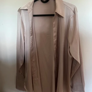 Beige satinliknande skjorta S - Snygg beige skjorta i satinliknande material med klassisk krage och knappar framtill. Skjortan har långa ärmar med manschett och en stilren, minimalistisk look. Perfekt för dig som gillar en clean och trendig stil.