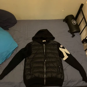 Svart Moncler pufferjacka med huva - Säljer en svart pufferjacka från Moncler Genius med huva och dragkedja. Jackan har stickade ärmar med en stor vit detalj och Moncler-logga på ena ärmen. Framsidan är quiltad och vadderad medan ryggen och ärmarna är i mjukt stickat material. Perfekt för dig som vill ha en snygg och unik jacka till höst och vår, priset är ej hugget i sten